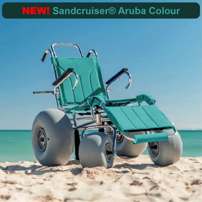 A blue and white beach wheelchair with large, wide wheels designed for smooth movement across sand. The wheelchair is placed on a sandy beach near the shoreline, with the ocean and a clear blue sky in the background.